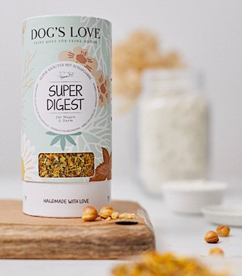 Auf dem Bild steht eine Packung DOG'S LOVE Super Digest Kräuter auf einem Holzbrett um diese liegen ein paar selbstgemacht Snacks auf einem weißen Untergrund.